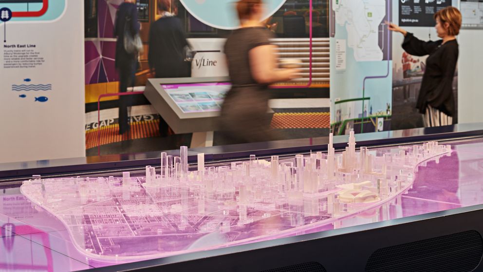 Platic model of Melbourne in the foreground with people walking past displays in the background inside the Metro Tunnel HQ.