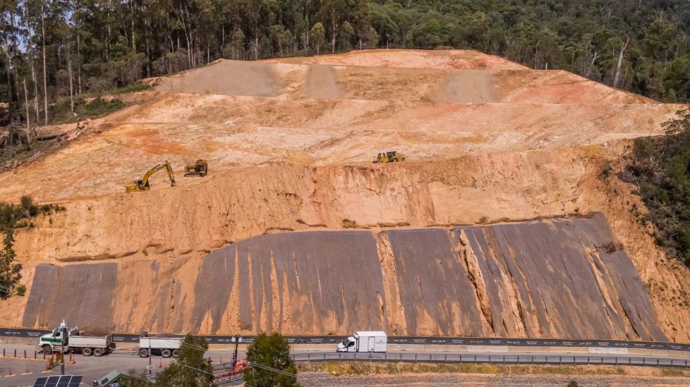 Trucks pass across the landslip on Bogong High Plains Road while machines continue to clear material above