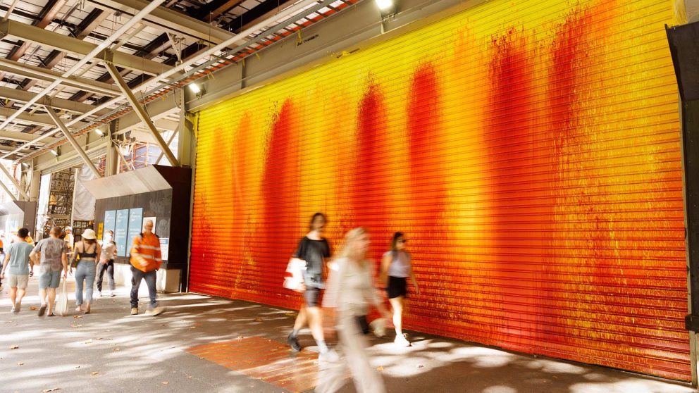 Pedestrians walking past a very large metal roller door, colourfully painted in red, orange and yellow. 