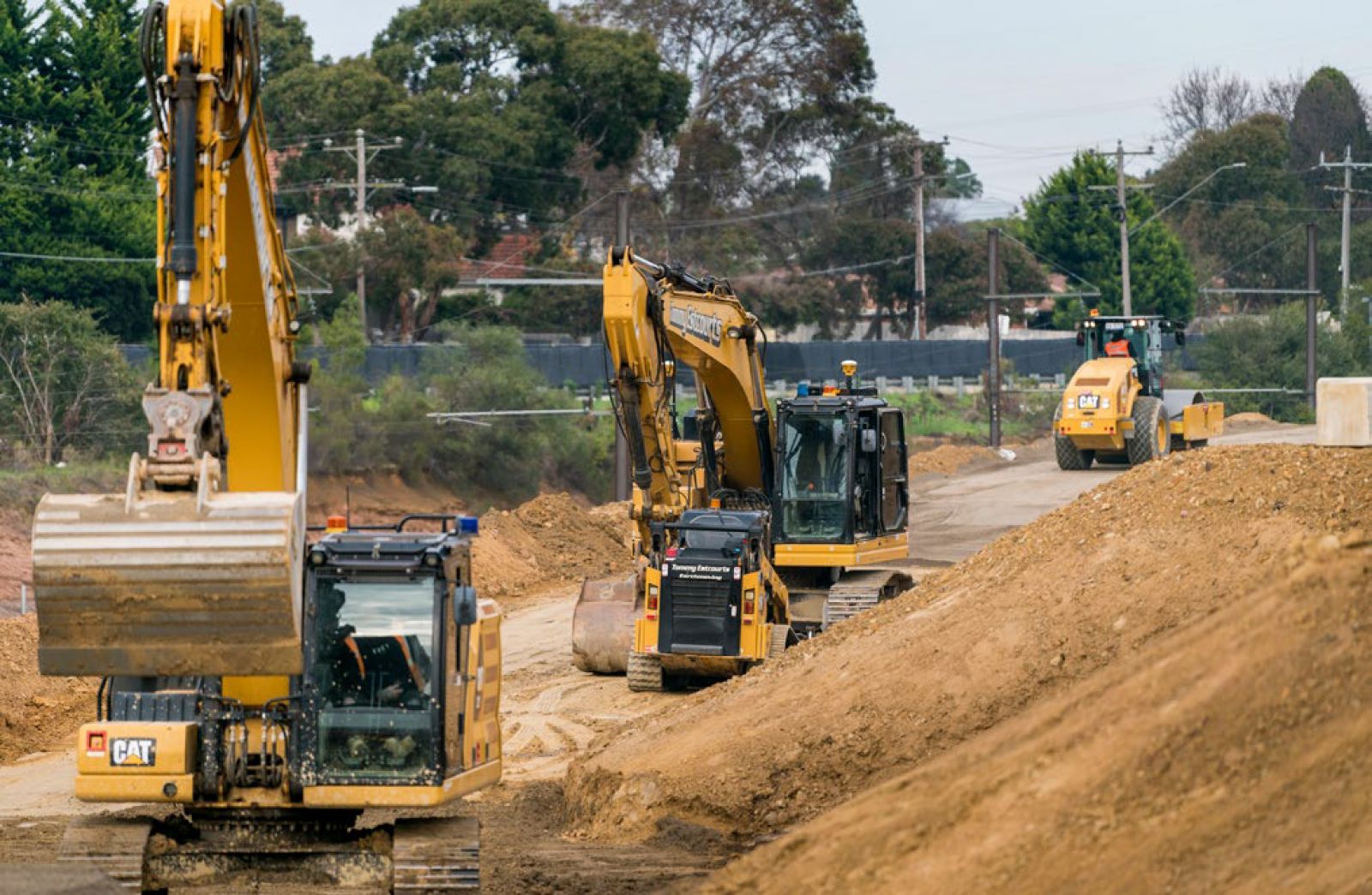 Construction machinery working on a dirt pathway, with excavators and a roller moving soil.