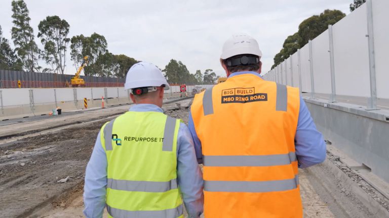 2 people in high vis and hard hats standing in the middle of a road under construction with machinery and trees in the background. Also visible is an electronic sign to show the speed limit of 60 km/h