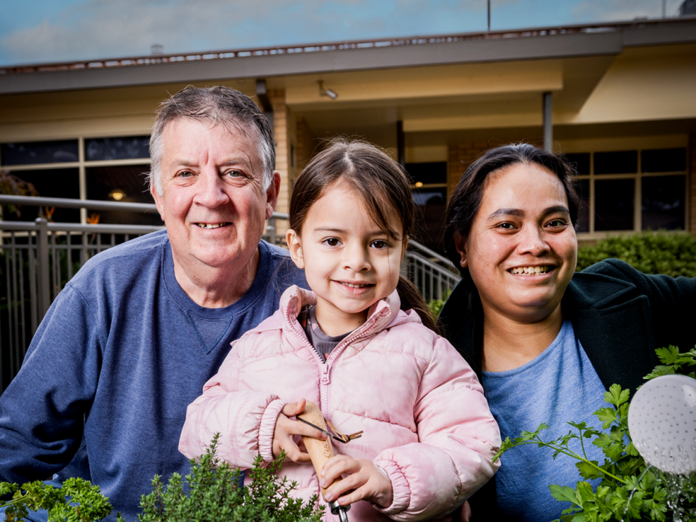 Image shows family of 3 smiling in the garden.