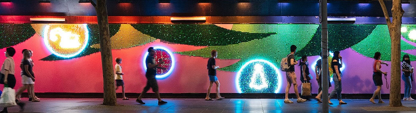 People walk past a neon-lit mural showing Christmas trees and digging machines.