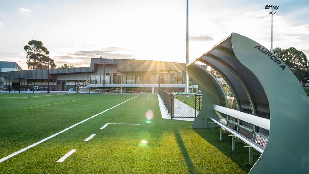 The sun setting on Greensborough College oval looking towards the newly built pavilion. There's a small covered grandstand in the foreground overlooking the new synthetic turf soccer pitch.  
