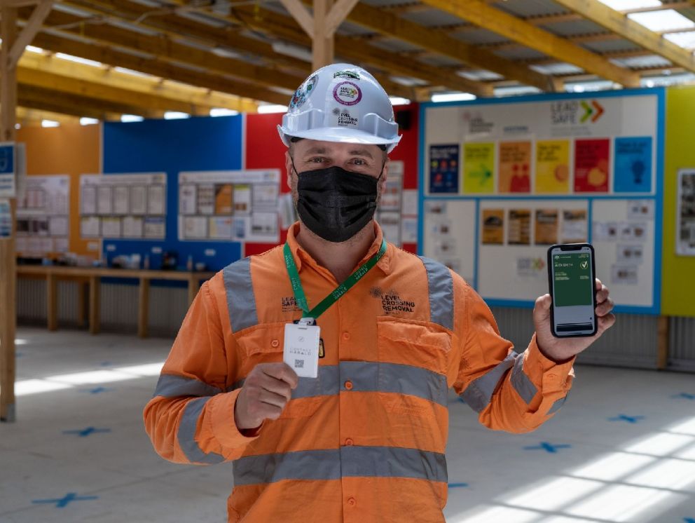 A man facing the camera smiling under his mask. He's holding up a phone which displays his vaccination status and the other hand holds a contact tracing card.