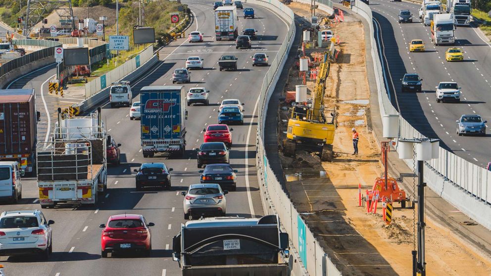 A work site in the centre of the West Gate Freeway with construction vehicles and workers with traffic travelling on the freeway on either side of the construction site.