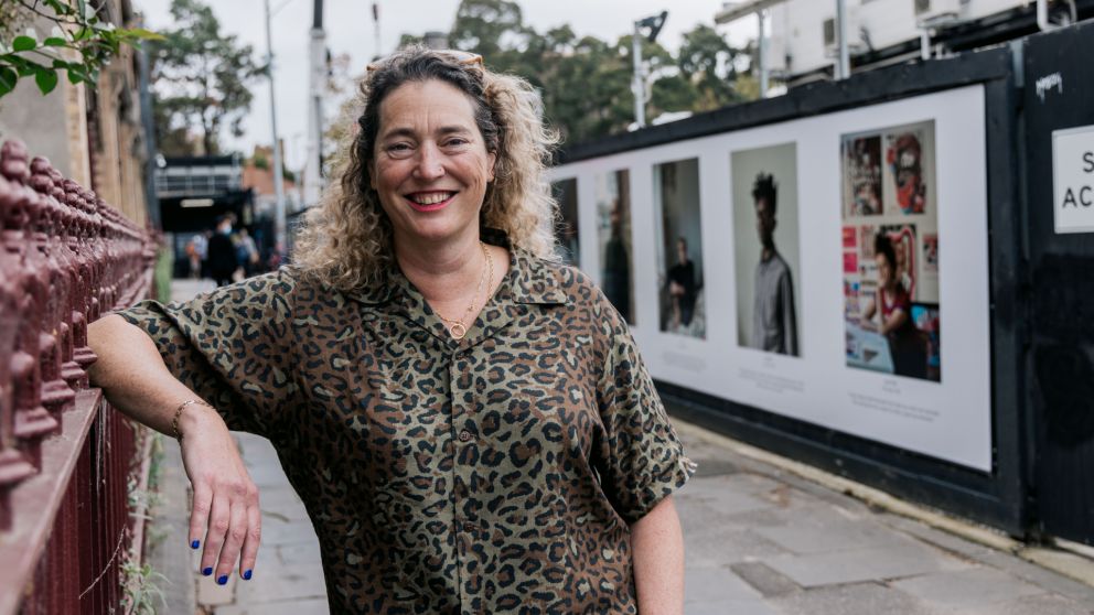 Jenny Lewis stands smiling in front of portraits on the hoarding for the One Hundred Years project