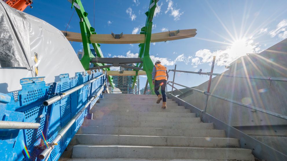 A worker in bright orange vest walks up a concrete staircase under the canopy legs and crossbeams.