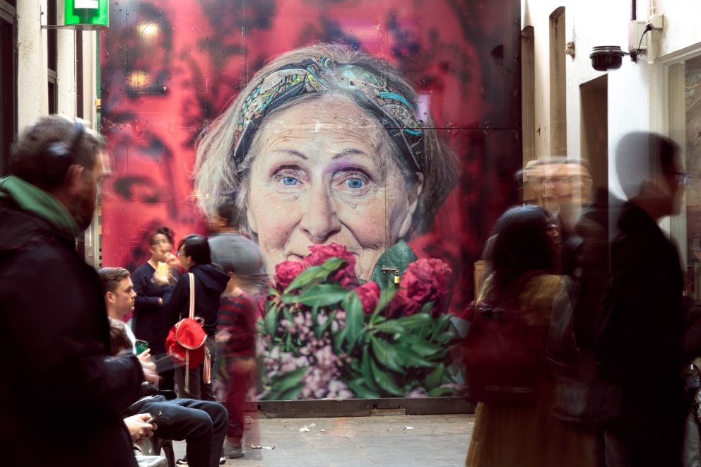 A bright artwork at the back of a bustling alleyway with a cafe, depicting an older woman's face on a pink background with flowers in the foreground