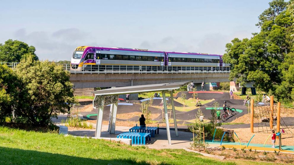 A hilly BMX track, next to a playground and covered sitting area. A V/Line train passes over an elevated bridge behind.