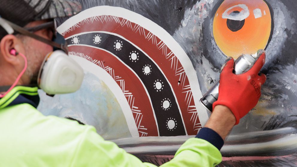 A man in a protective mask spray paints a section of an artwork on a wall