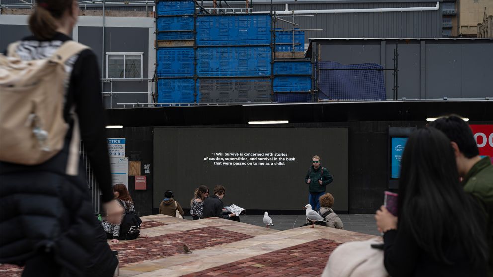 Image of the Fed Square with people sitting on the steps. In the background is the Town Hall construction site with two screens installed on the construction hoarding displaying two black and white photographs. 