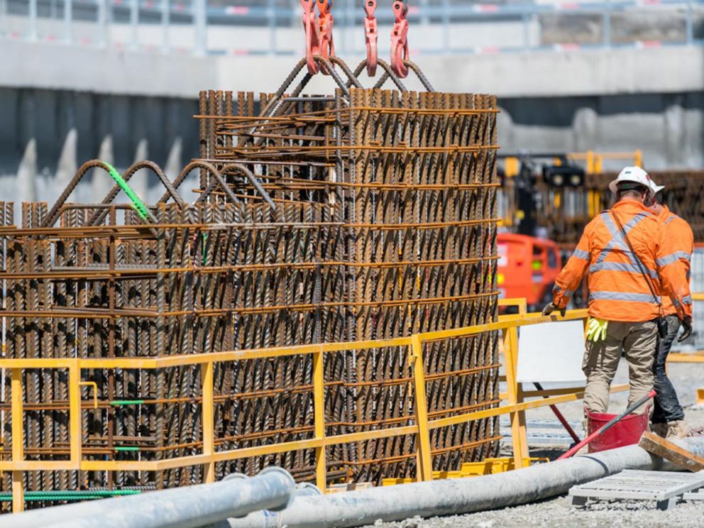 Two construction workers in high-vis protective gear standing next to steel reinforcement cages being lowered into a D-wall on a North East Link construction site.