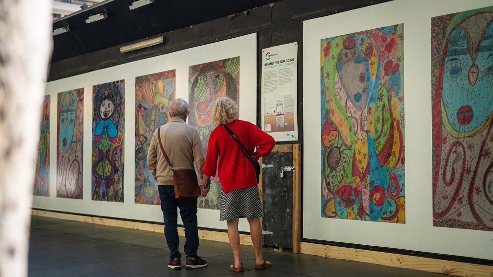 2 people standing in front of colourful construction hoarding, featuring a row of abstract portraits reading panel
