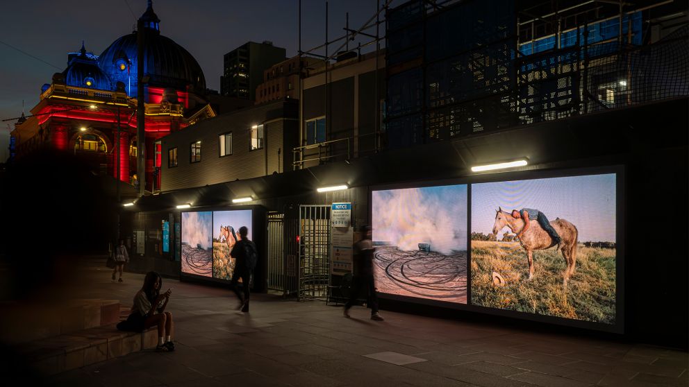 Pedestrians walking past 2 digital screens with images of the outback, placed on hoardings infront of a construction site