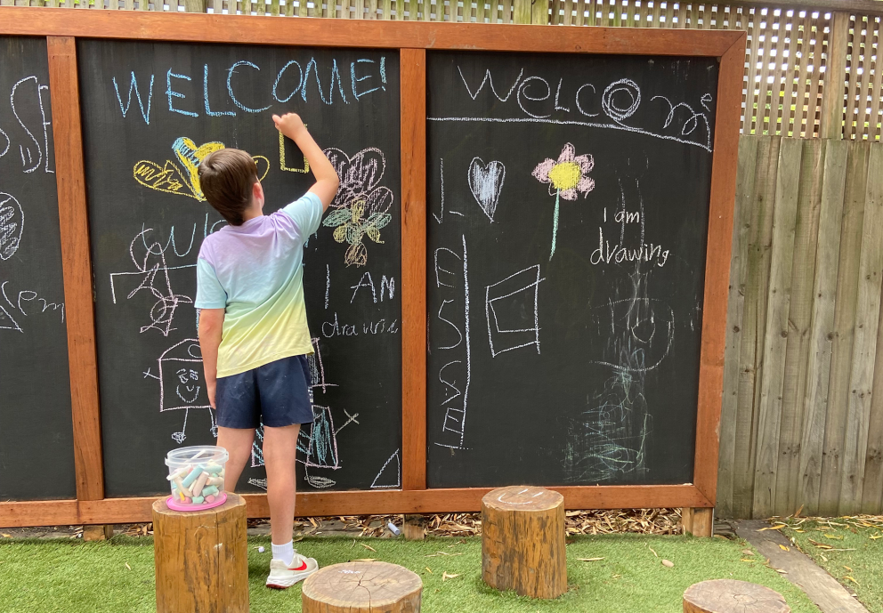 A child uses colored chalk to draw on an outdoor blackboard decorated with welcome messages and drawings. They stand on artificial turf next to a line of wooden stumps, one of which holds a tub of chalk.