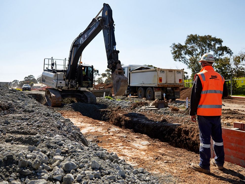 Image of of an excavator creating a trench with a construction worker watching the process.