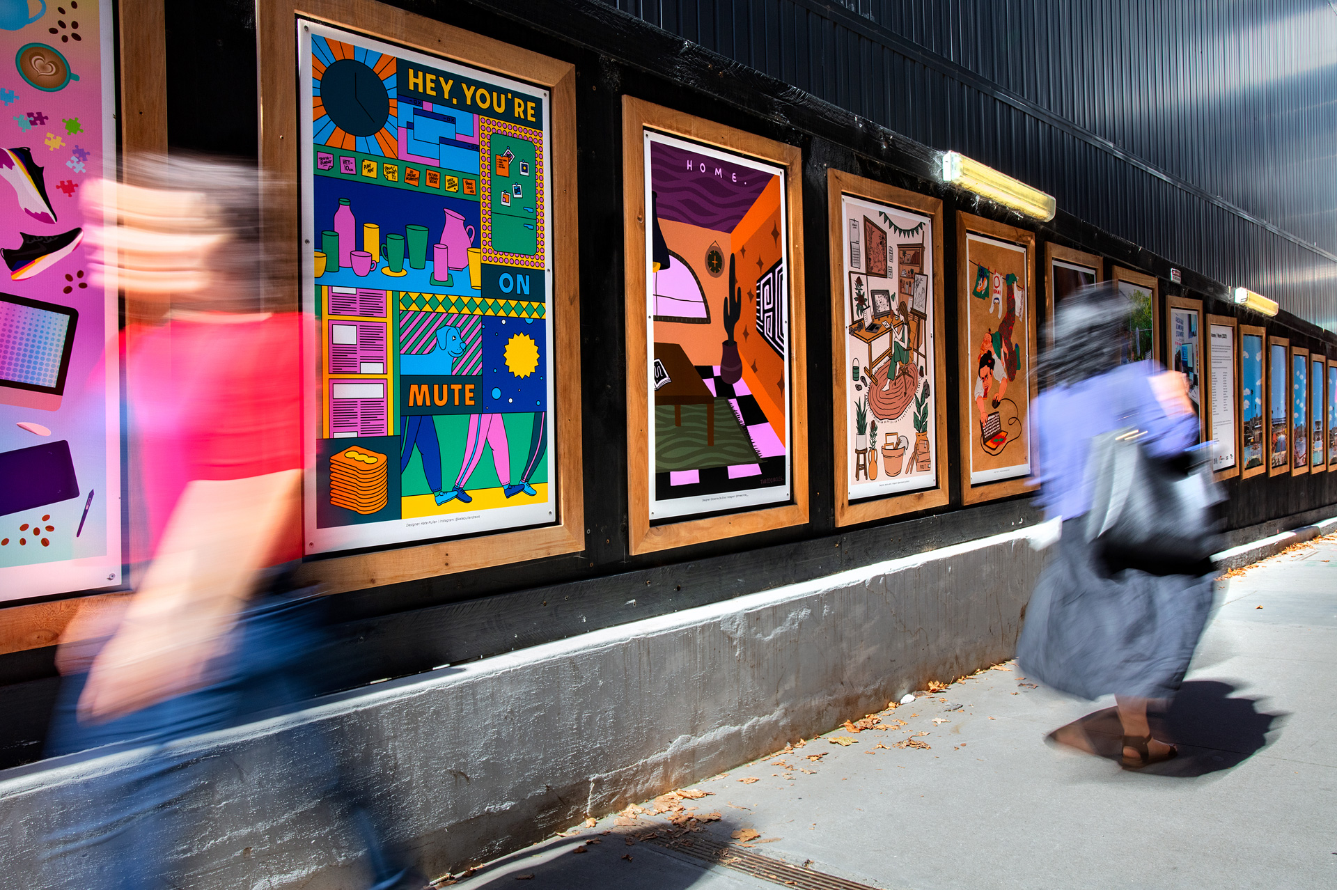 Two blurry people walk past brightly coloured framed artworks on a street