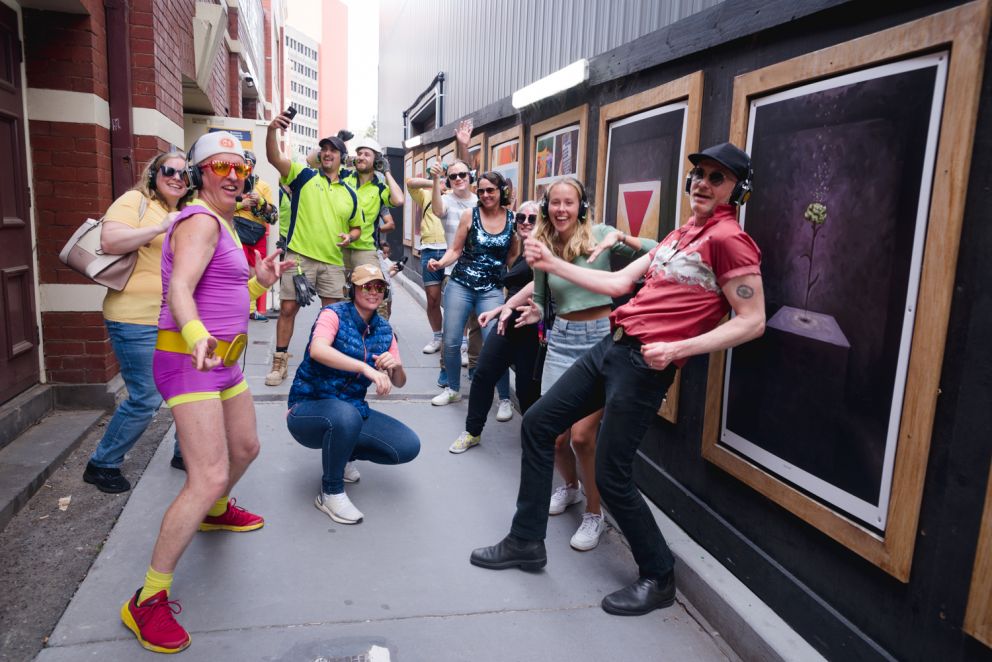 People dressed in colourful outfits wearing headphones dance next to artworks in a laneway