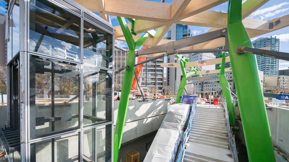 A view looking towards the tram interchange staircase, lift shaft and pedestrian bridge under the green legs and wooden crossbeams of the canopy.