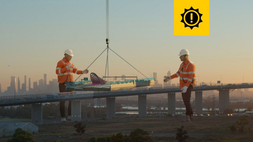 Two giant-sized men in hard hats and work wear stand next to the West Gate Bridge, helping to manoeuvre a segment of road with a tunnel entrance, which is suspended from a crane. 