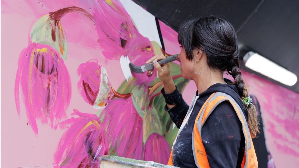 A woman paints large flowers on a wall.