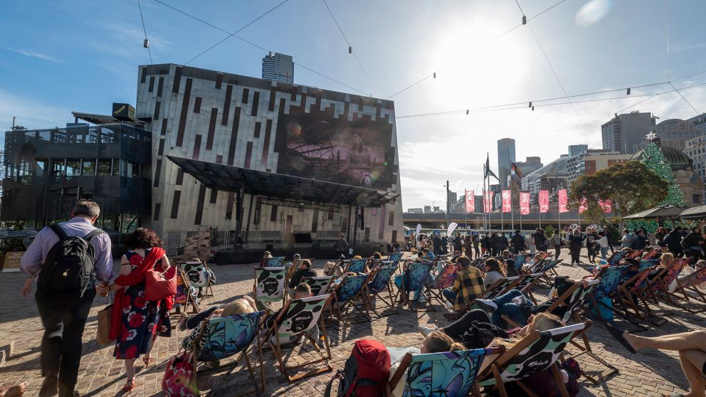 A large number of people sit in chairs in the sun at Fed Square