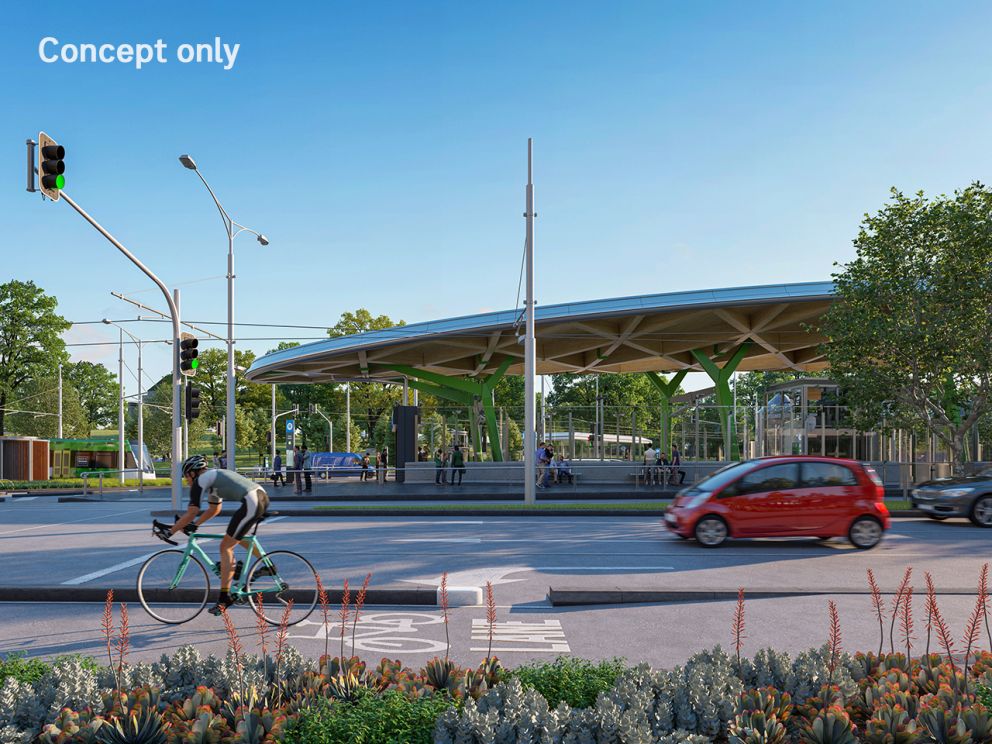A render of Anzac Station entrance exterior from Albert Road. The entrance canopy is in full view with blue skies in the background. A bike is riding past in the bike lane and cars are driving by too.