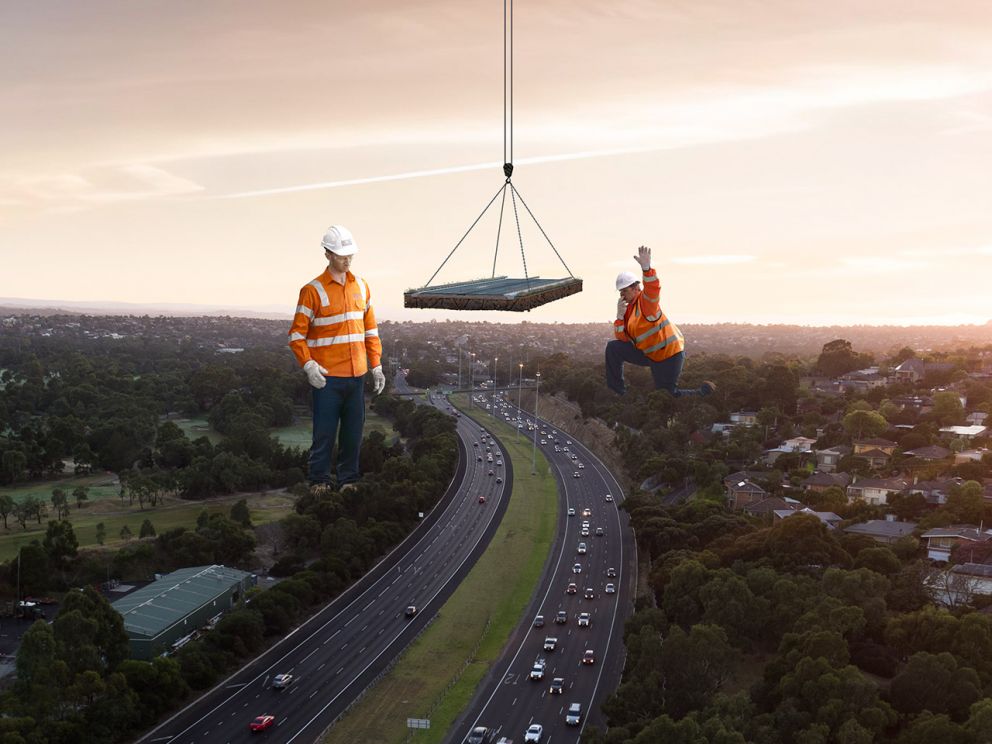 Two giant construction workers standing over a busy freeway with a section of road being lowered down from a crane