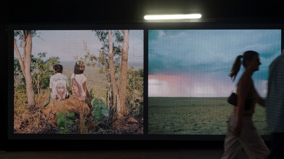 A figure walks past 2 digital screens with images of the outback