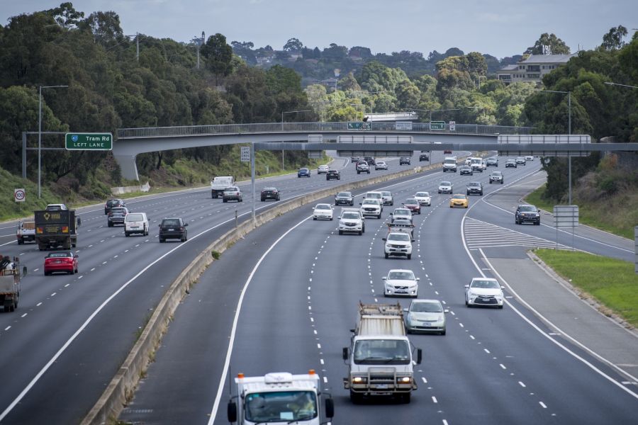 An elevated view of a busy highway with cars and trucks traveling in both directions. The road passes under a concrete overpass, with signs pointing to 