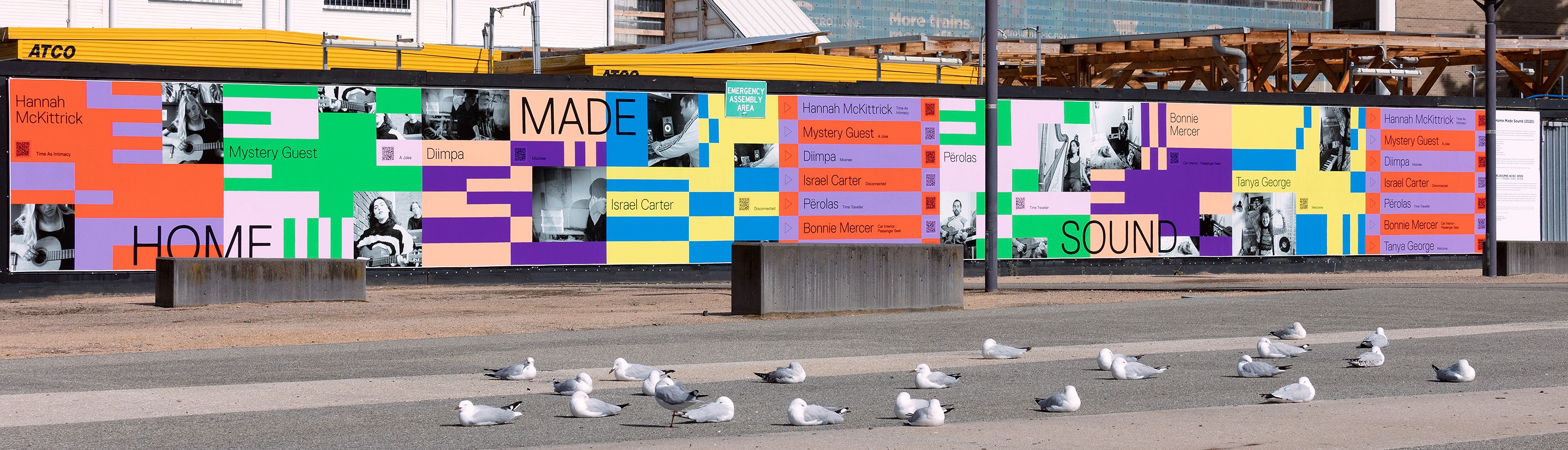 A mural featuring images of musicians and blocks of colours covers construction hoarding in a plaza.