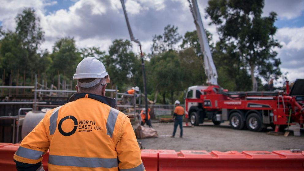 A construction worker in high-vis clothing and hard hat standing onsite at Trinity College. He stands in the foreground with his back to camera watching other construction workers onsite in the background near a truck. 