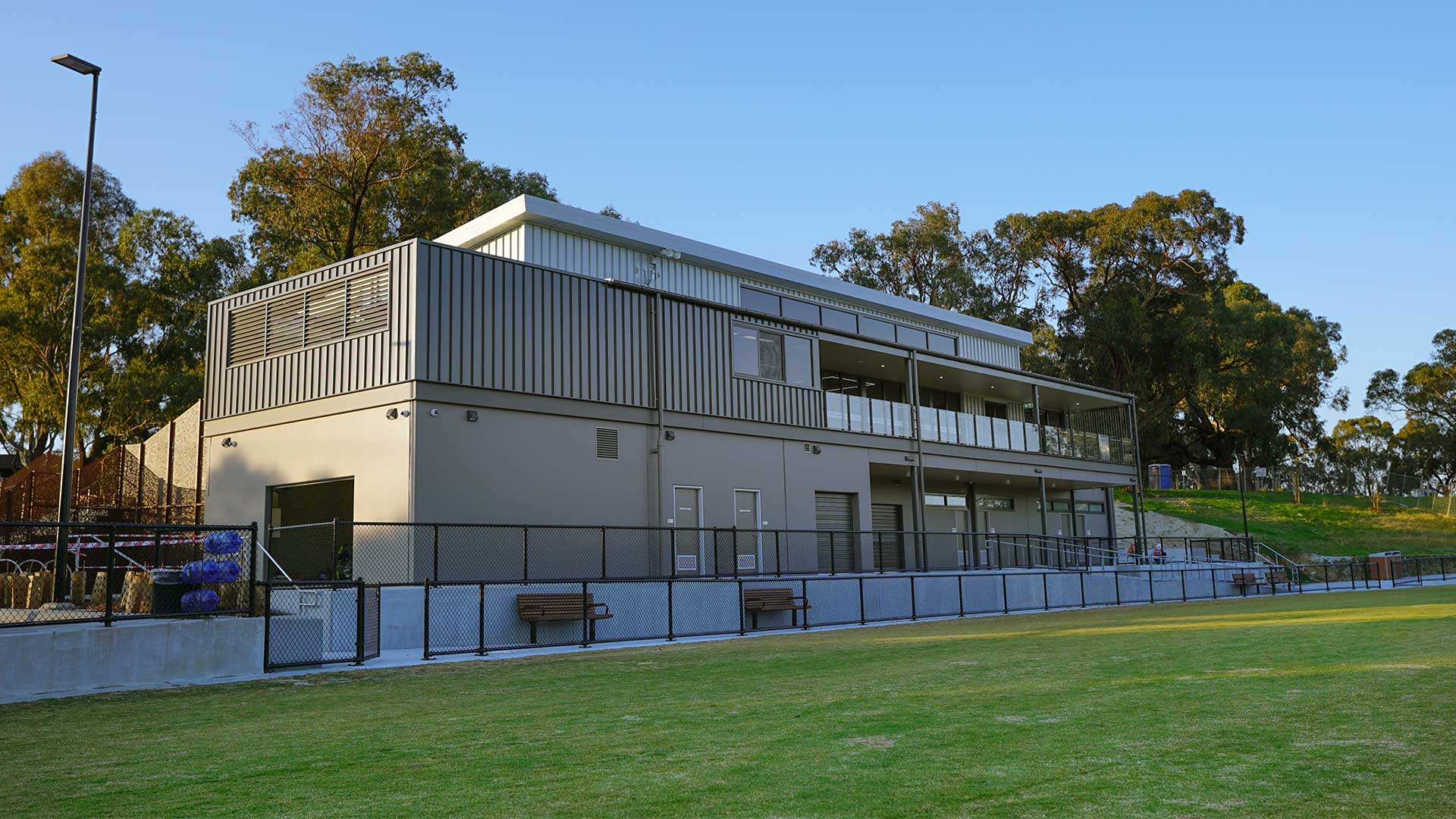 Looking at Binnak Park pavilion from the oval. The pavilion has been built on a grassed embankment which leads up to a line of mature trees behind the pavilion. 