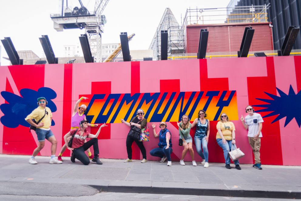People dressed in colourful outfits wearing headphones dance next to a mural that says 'Community'