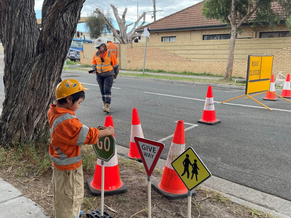 Small boy in orange work wear holding a traffic sign near a road closure