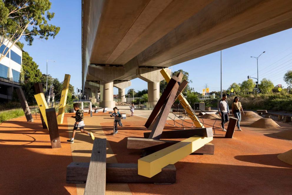 Several children play in a paved playground area underneath an elevated train line. The equipment is made from large square-cut logs.