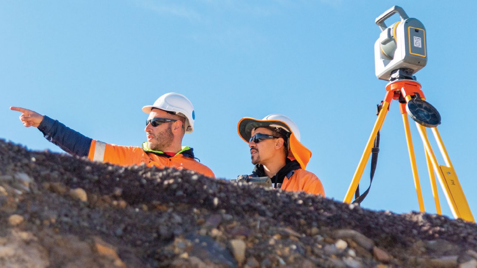 Two construction workers wearing high-vis protective gear next to surveying equipment.