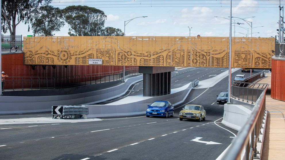 A rail bridge over a busy road. The rail bridge has decorative designs pressed out of pieces of metal that line the bridge.