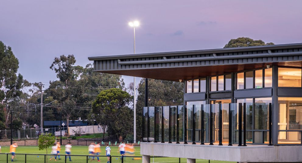 A soccer team practicing on the oval at dusk in front of the Greensborough College pavilion. The floodlight on the oval is turned on and the lights are on inside the pavilion.