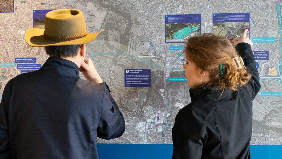A man and woman viewing a large map on a wall. The man is wearing a wide brim hat and dark jacket and the woman is pointing to an image on the map.