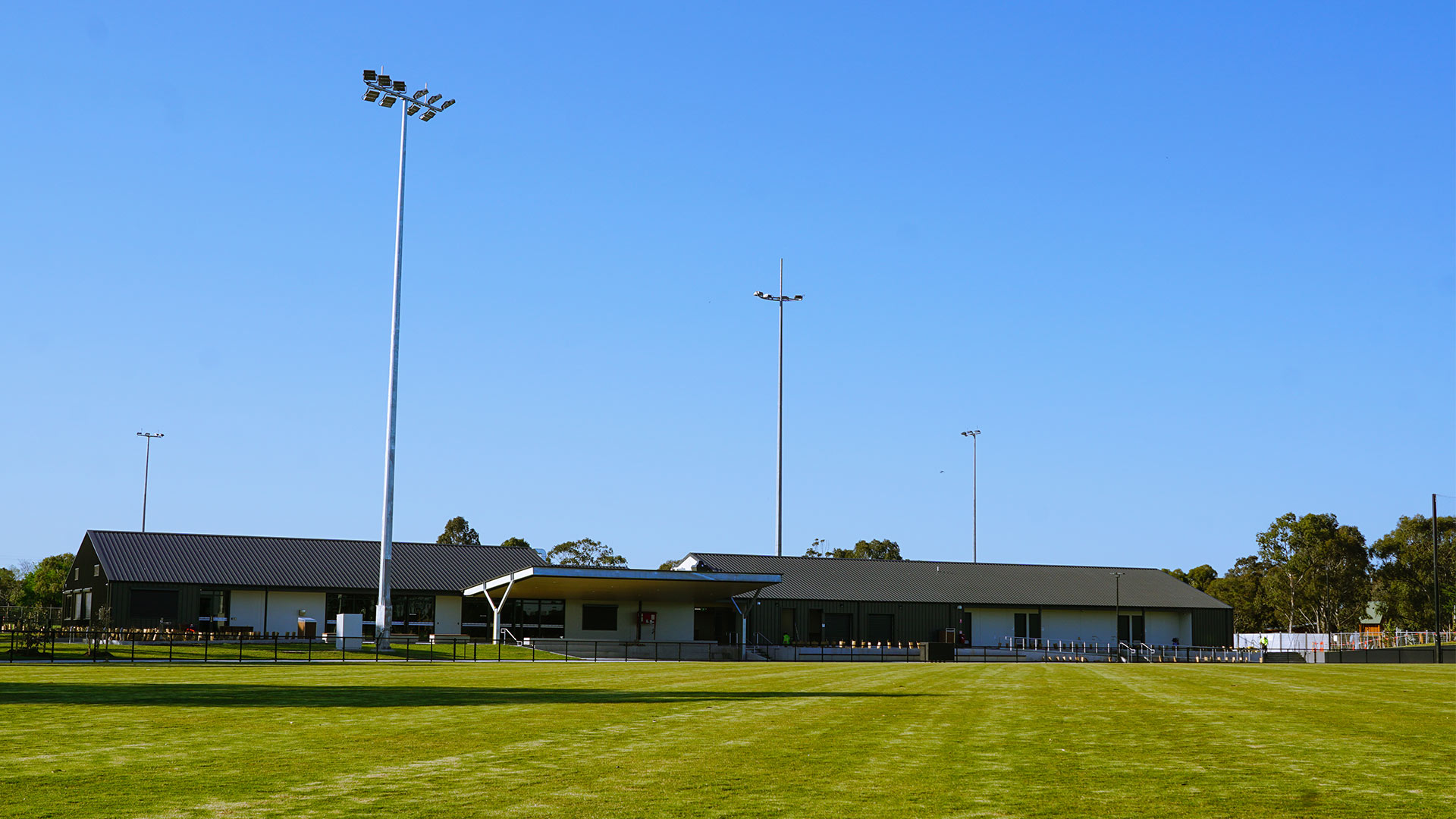 Looking at Ford Park pavilion from the oval. Mature trees are behind the pavilion and there are four floodlights positioned around the oval.