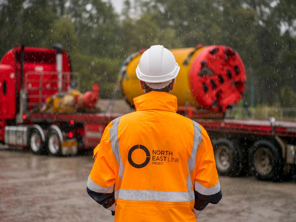 A construction worker in high-vis clothing and hard hat standing in the rain. He stands in the foreground with his back to camera looking at a mini tunnel boring machine on the back of an oversized truck.