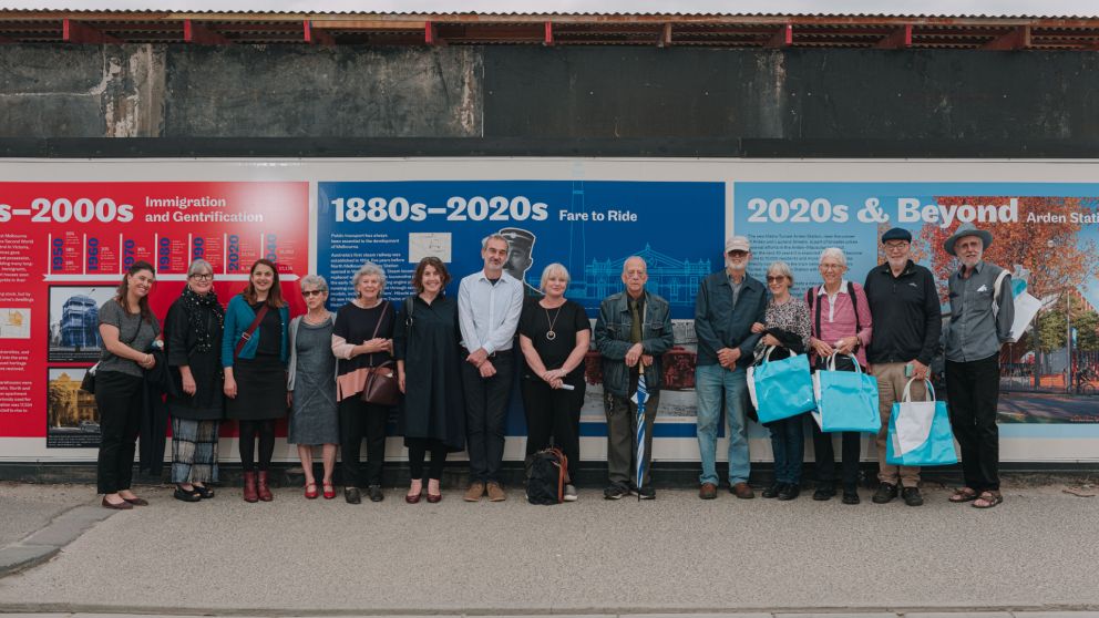 14 people stand in front of the historical hoarding artwork at Arden Station