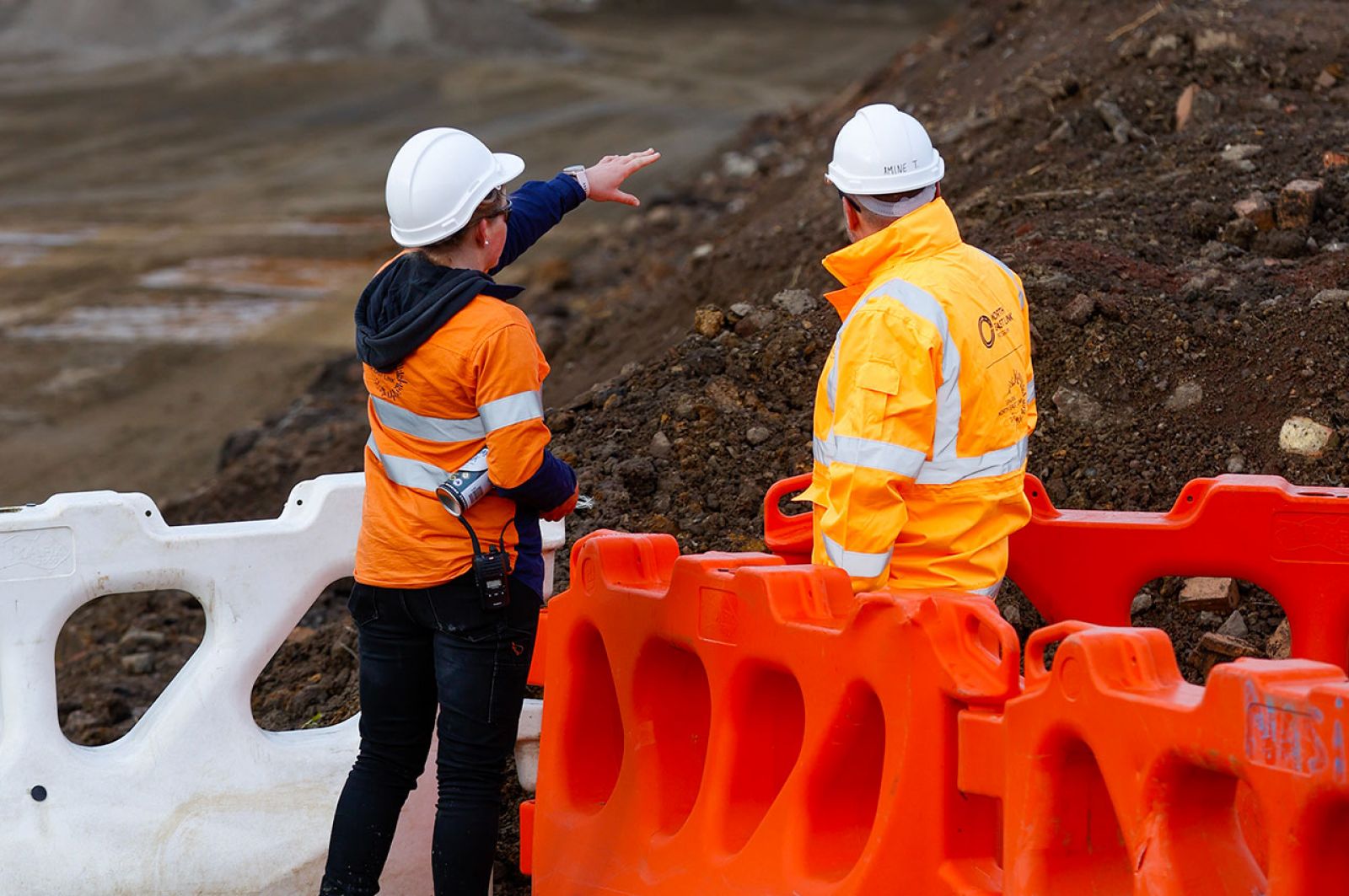 Two construction workers wearing high-vis and protective equipment standing on a worksite. One of the workers is pointing to a large mound of soil. 