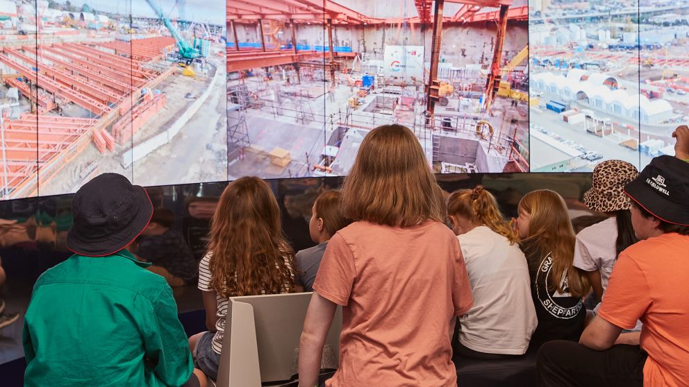 Group of people sitting in the foreground watching a panel of digital screens of construction in the background.