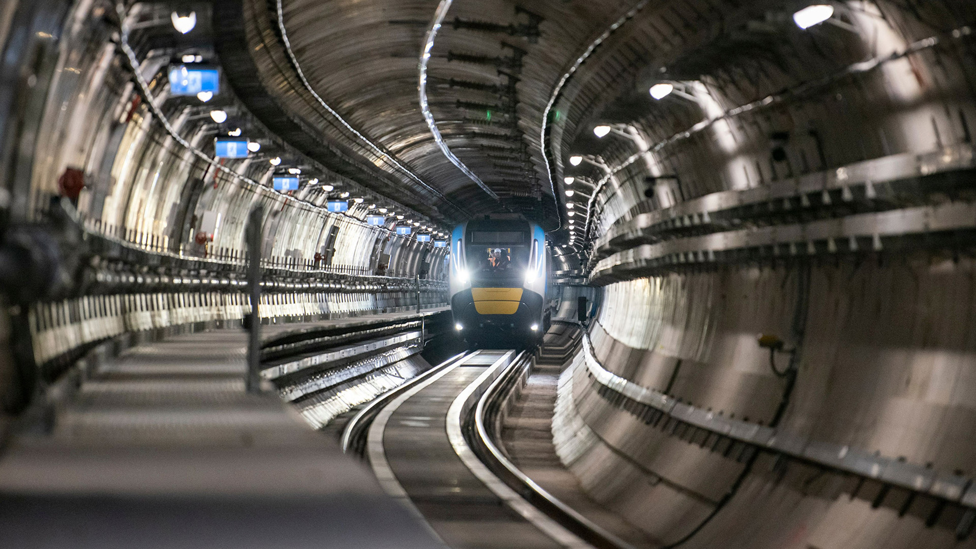 The front of a blue and yellow train can be seen inside a concrete train tunnel.