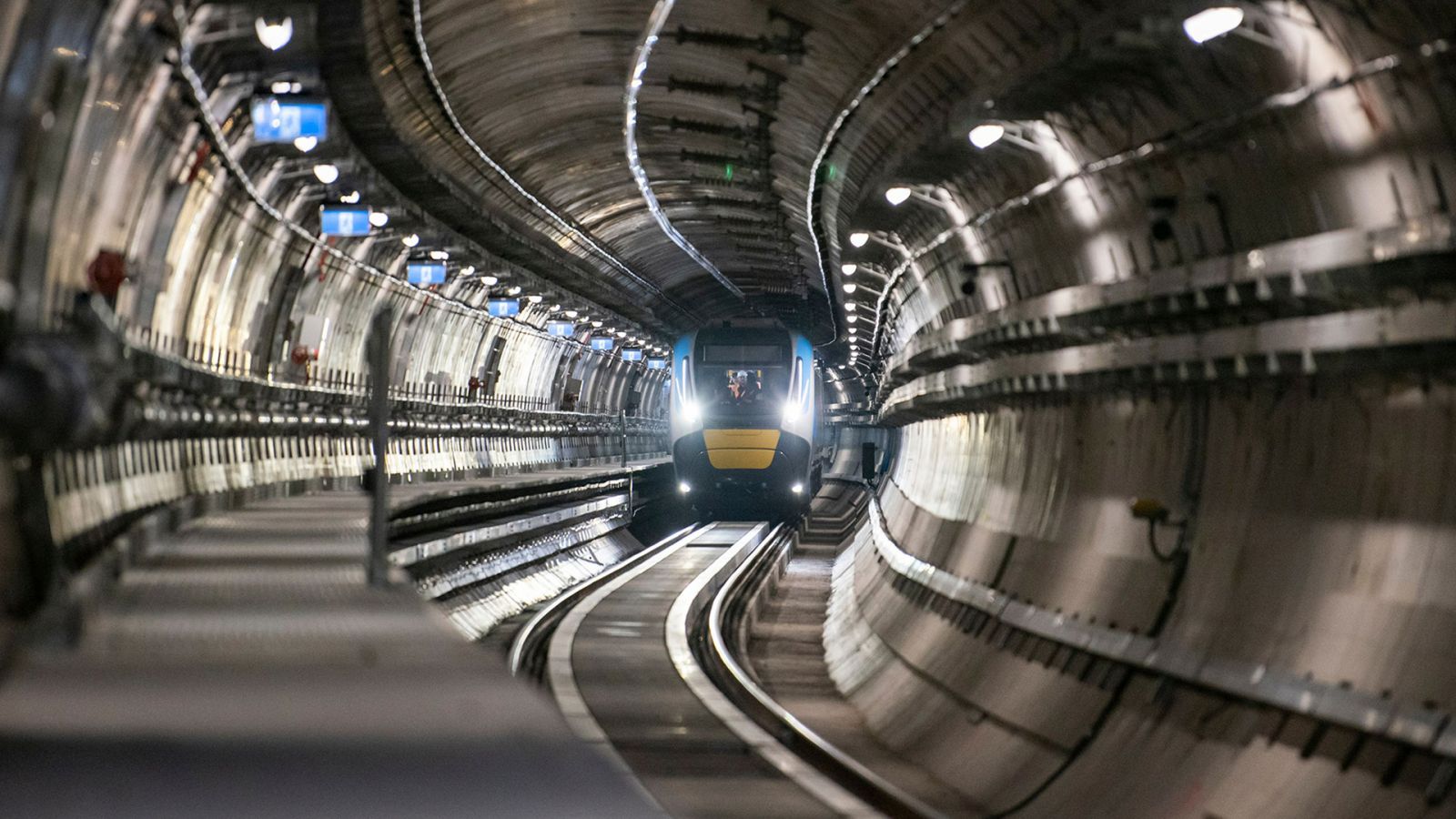 The front of a blue and yellow train can be seen inside a concrete train tunnel.