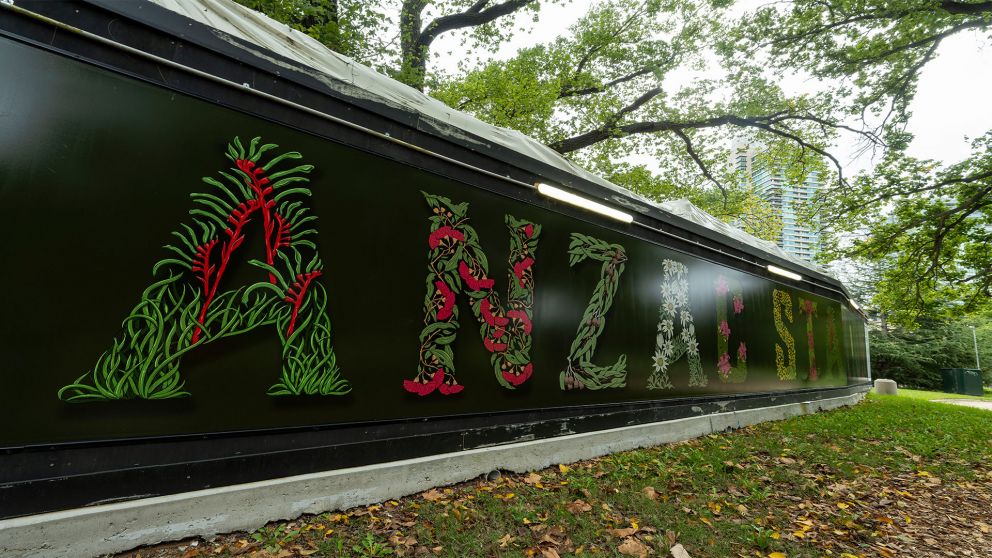 A photo of a construction hoarding with floral letters, in front of fallen leaves on grass, with leafy green trees in the background.