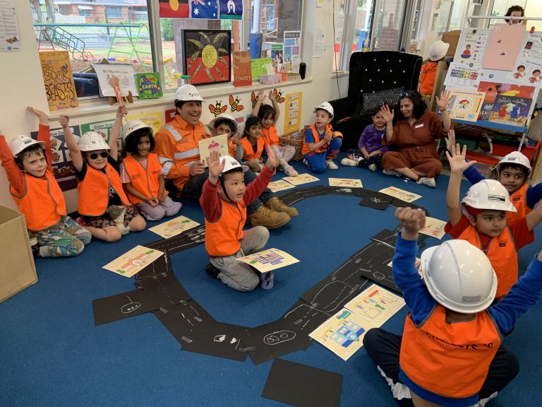 Kinder class with kids wearing high vis vests and hard hats. Kids are gathered around drawings in the middle. 